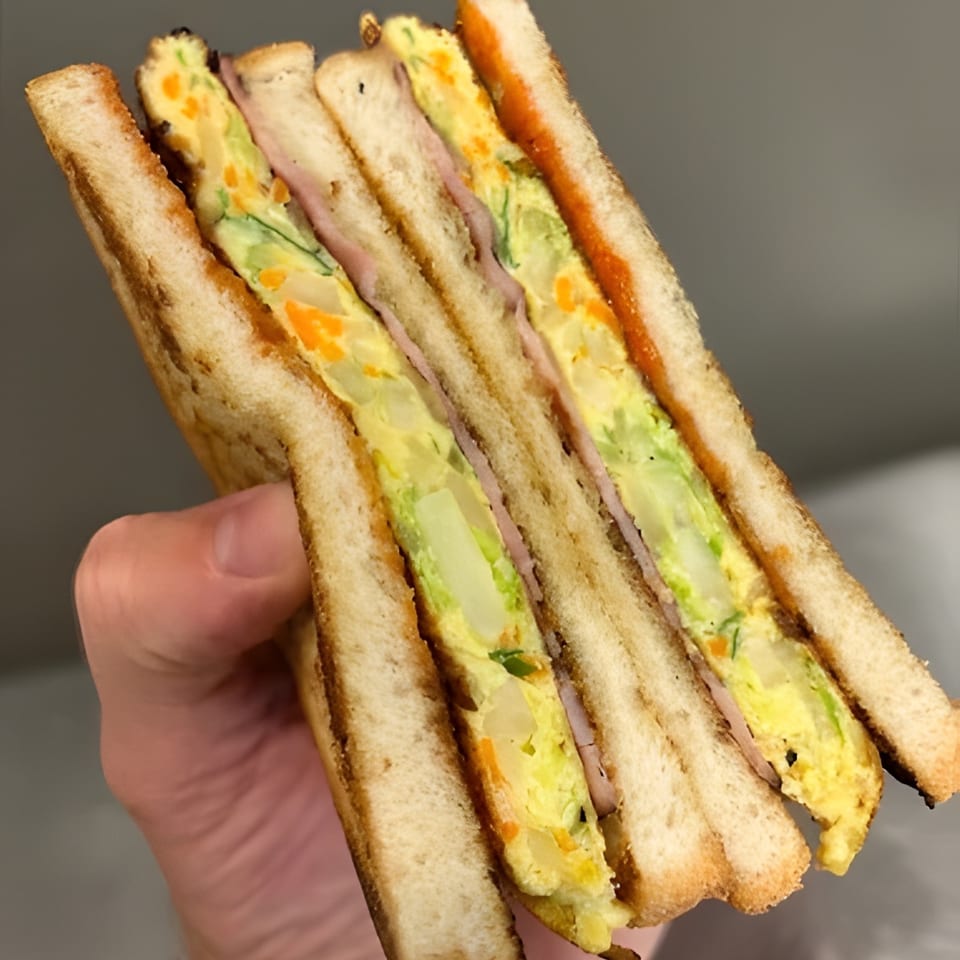 Hand holding a thick, layered sandwich with egg, ham, and vegetables on toasted bread.