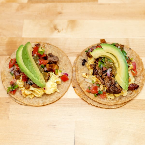 Two breakfast tacos topped with scrambled eggs, grilled meat, salsa, vegetables, and avocado slices on a wooden table.