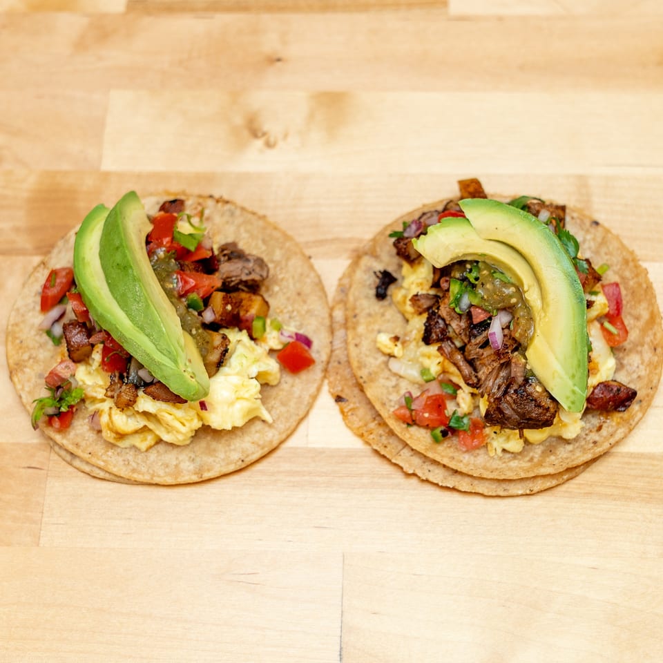 Two breakfast tacos topped with scrambled eggs, grilled meat, salsa, vegetables, and avocado slices on a wooden table.