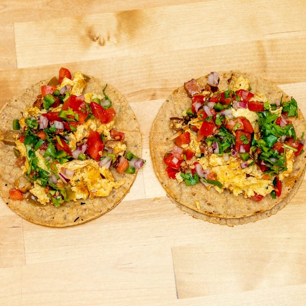 Two corn tortillas topped with scrambled eggs, diced tomatoes, peppers, onions, cilantro, and savory fillings on a wooden table.