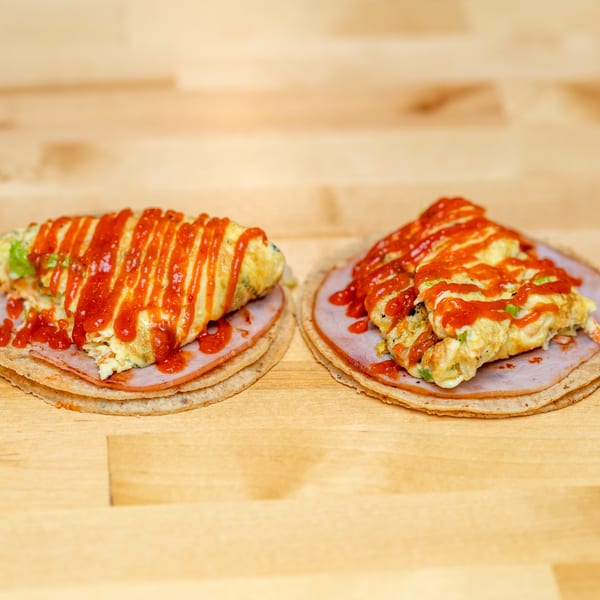 Two open-faced tortillas topped with sliced meat, vegetable-filled omelets, and bright red sauce on a wooden table.