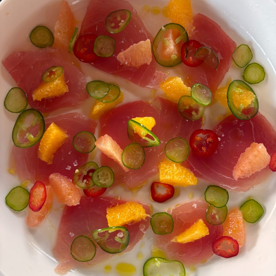 Thinly sliced raw fish with grapefruit, orange, cucumber, and red and green chili peppers on a white plate.