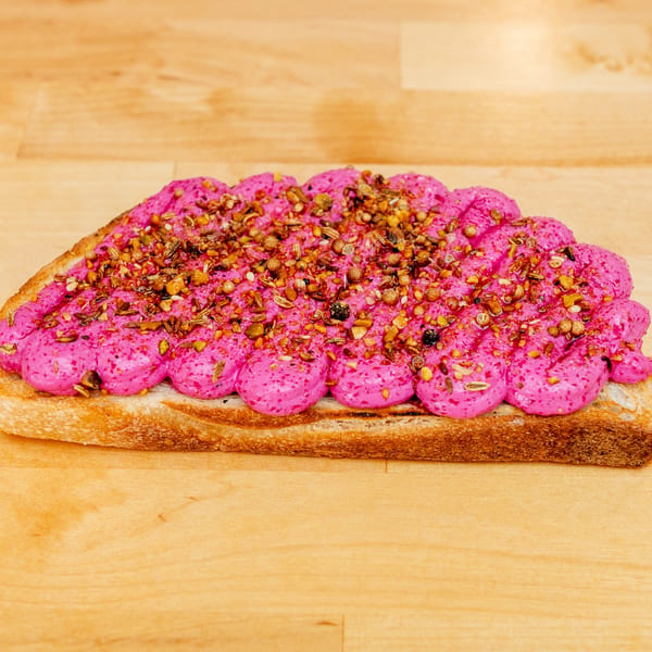 Thick slice of toast topped with bright pink spread and a generous sprinkling of toasted seeds and spices on a wooden board.
