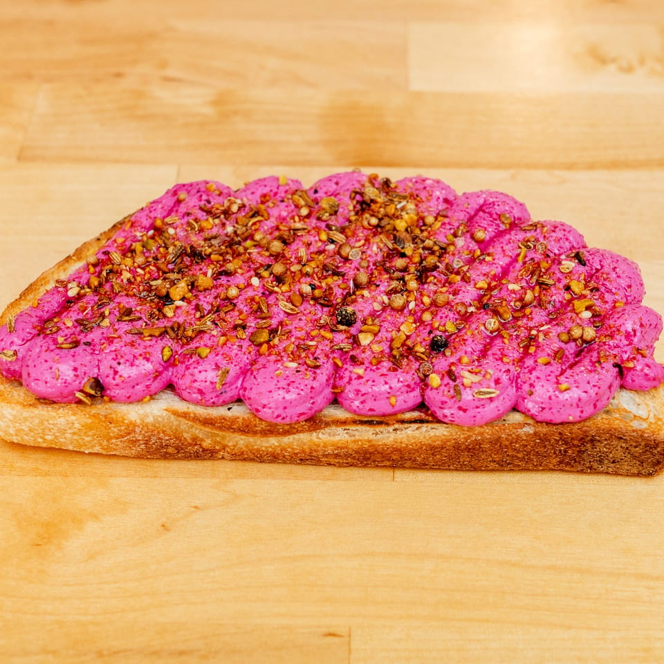 Thick slice of toast topped with bright pink spread and a generous sprinkling of toasted seeds and spices on a wooden board.