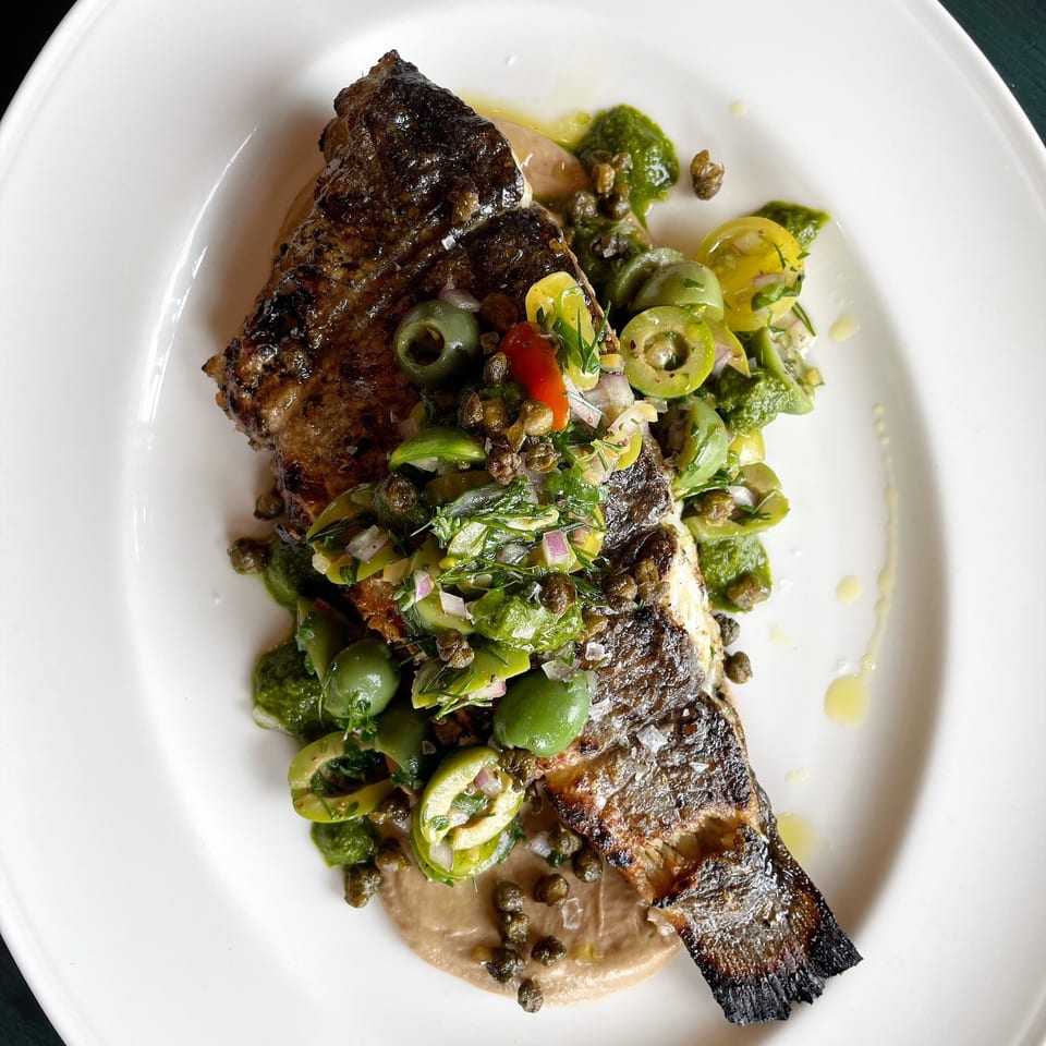 Crispy-skinned grilled fish topped with green olives, tomatoes, herbs, and capers over creamy sauce on a white plate.