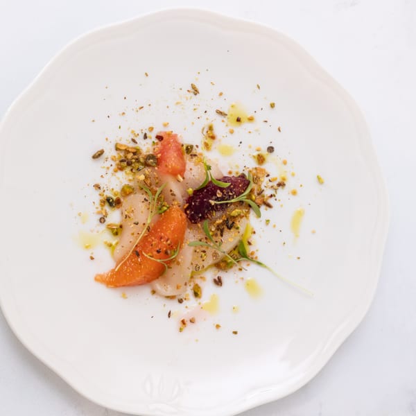 Delicately plated seafood with citrus segments, beet, pistachios, microgreens, and olive oil on a white plate.