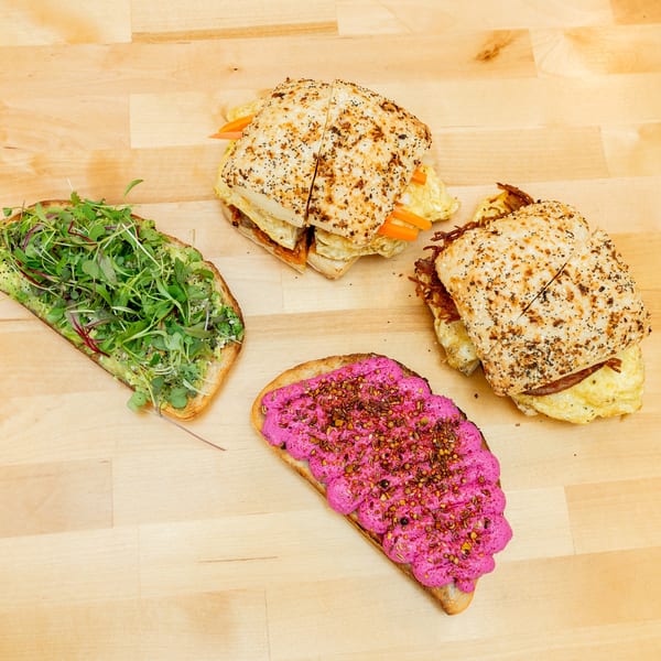 Assorted open-faced sandwiches on a wooden surface, including avocado with microgreens, seeded sandwiches, and bright pink beet spread.