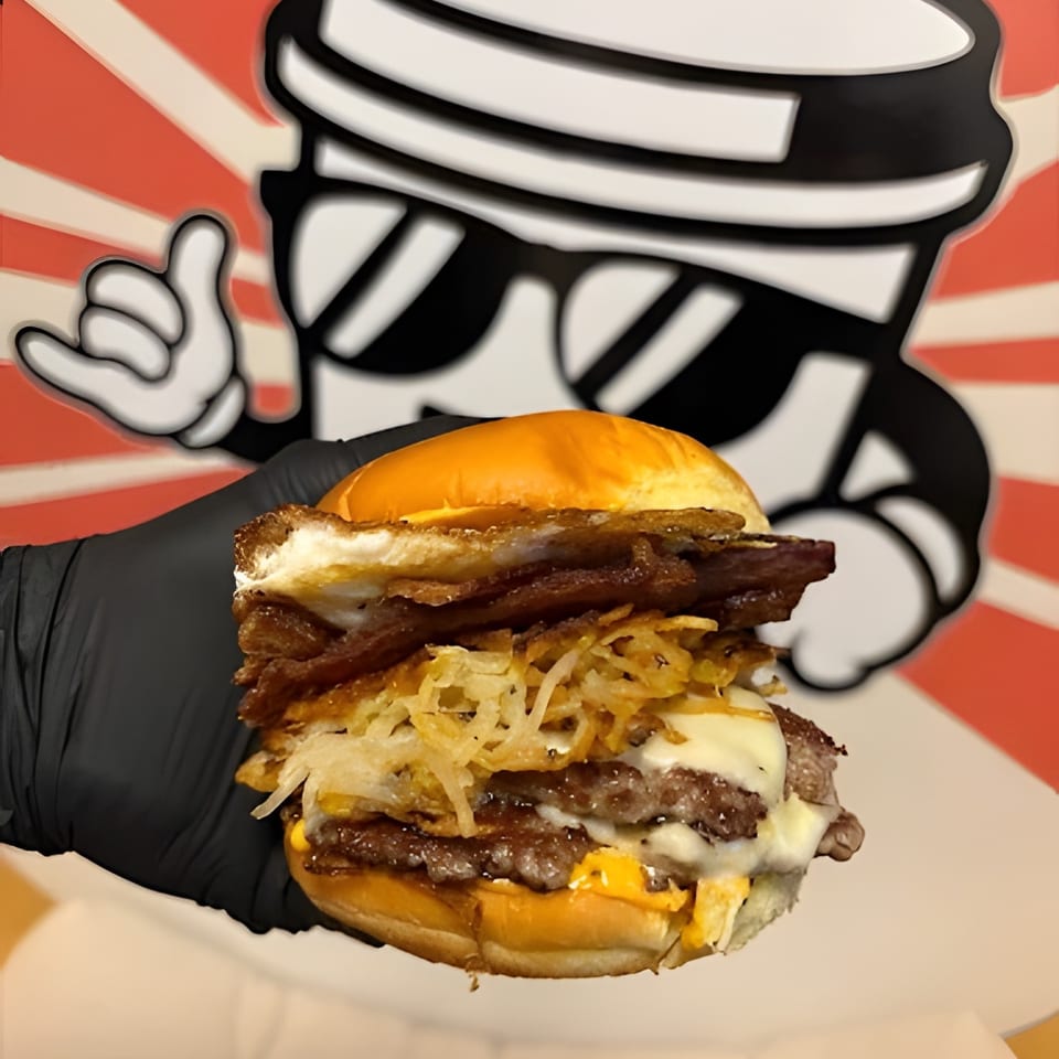 Gloved hand holding a breakfast burger with bacon, hash browns, egg, and cheese.