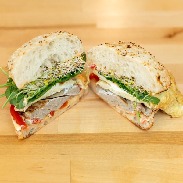 Two toasted sandwiches filled with sliced meat, cheese, spinach, sprouts, tomatoes, and creamy sauce on a wooden table.