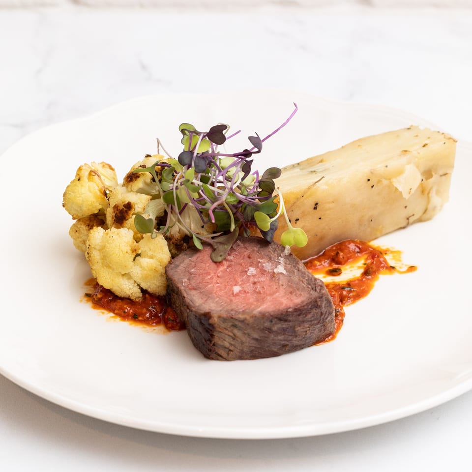 Seared beef tenderloin with roasted cauliflower, creamy potato, microgreens, and red pepper sauce on a white plate.