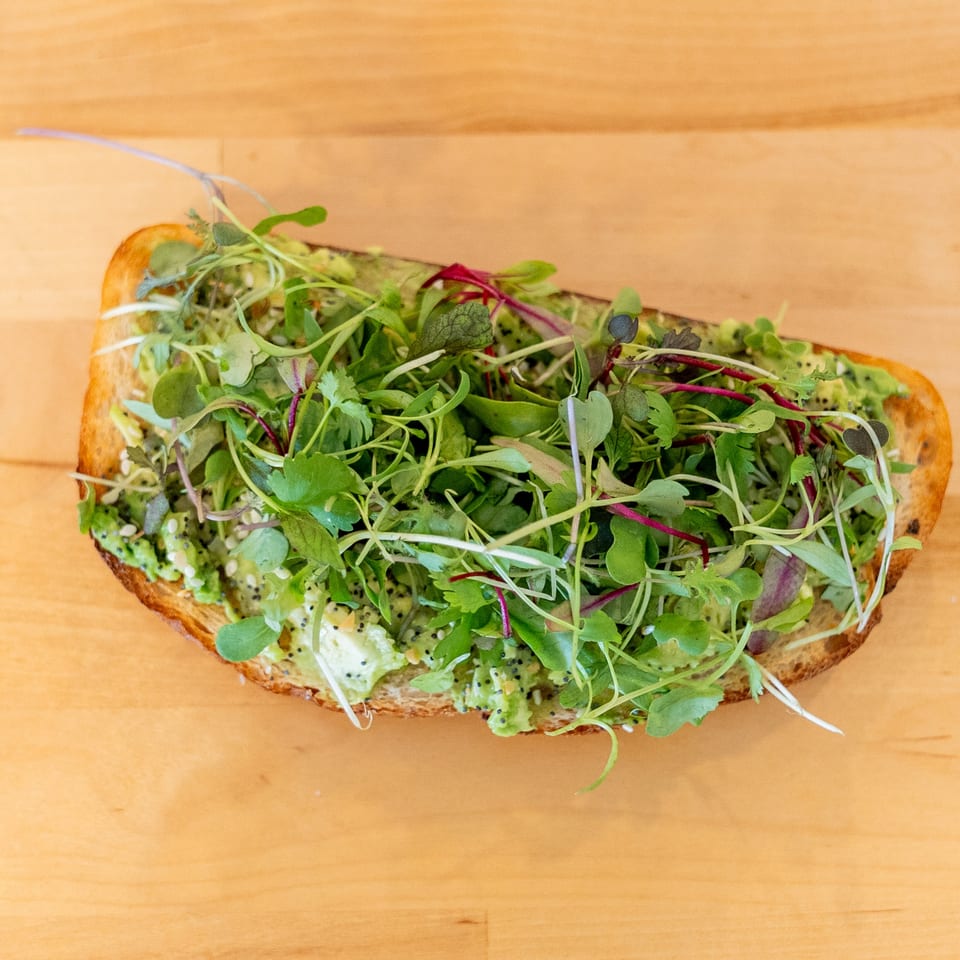 Open-faced toast topped with creamy spread, kiwi slices, sesame seeds, and abundant fresh microgreens on a wooden table.